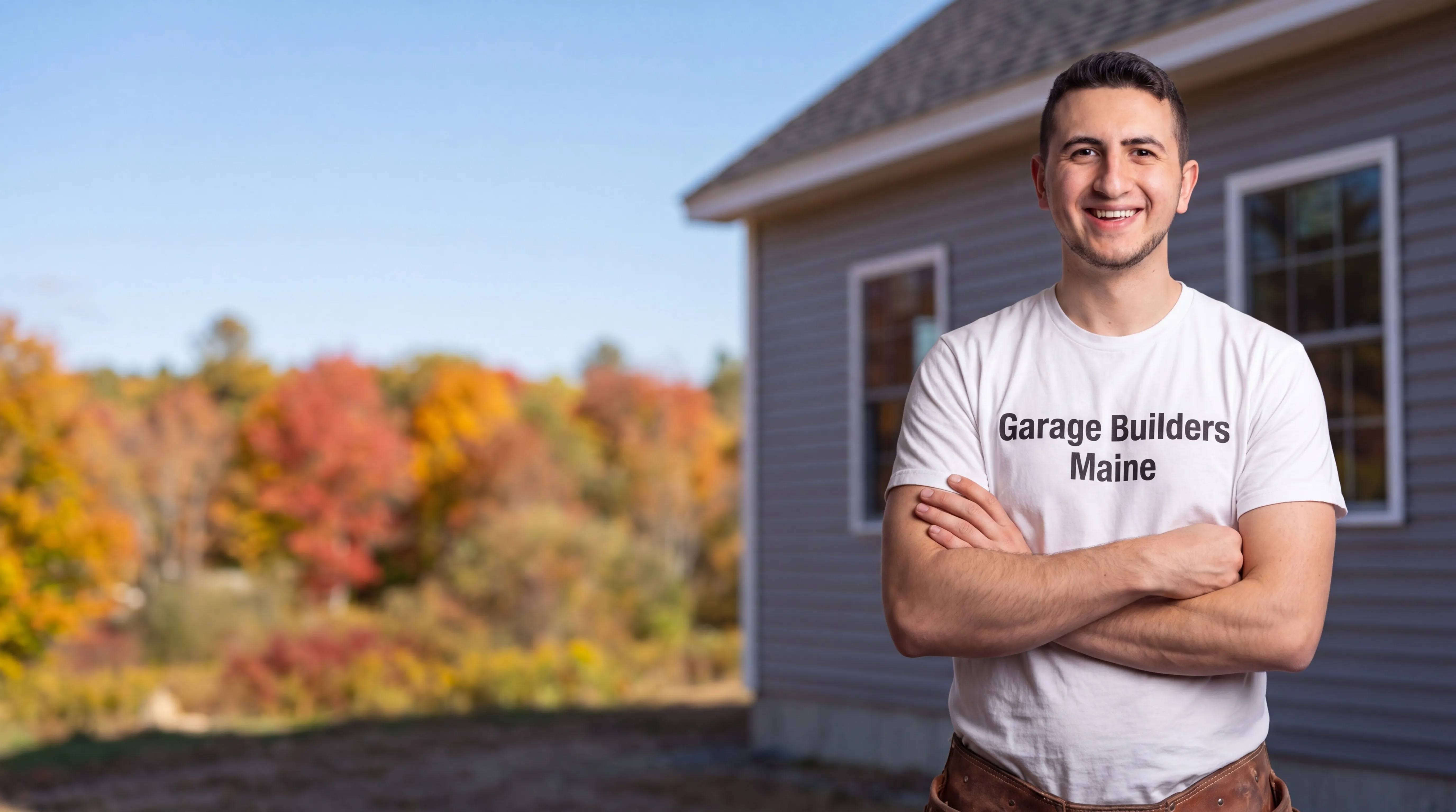 Garage Builders Maine owner with arms crossed beside the gray wall of a finished house in fall in Windsor, Maine