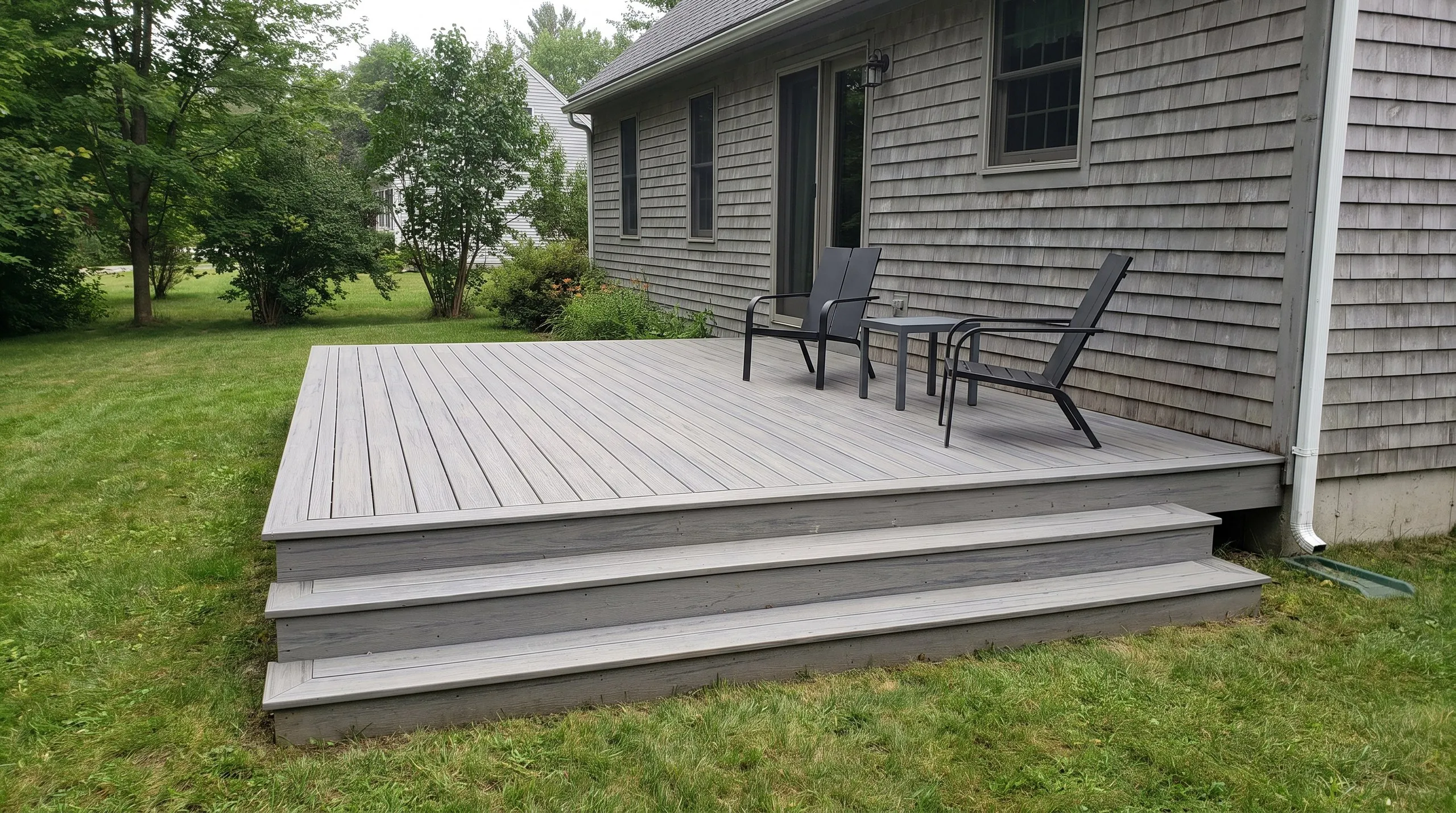 Deck contractor Winthrop Maine: low ground-level gray composite deck with two wide steps, two chairs and a side table by a shingled house